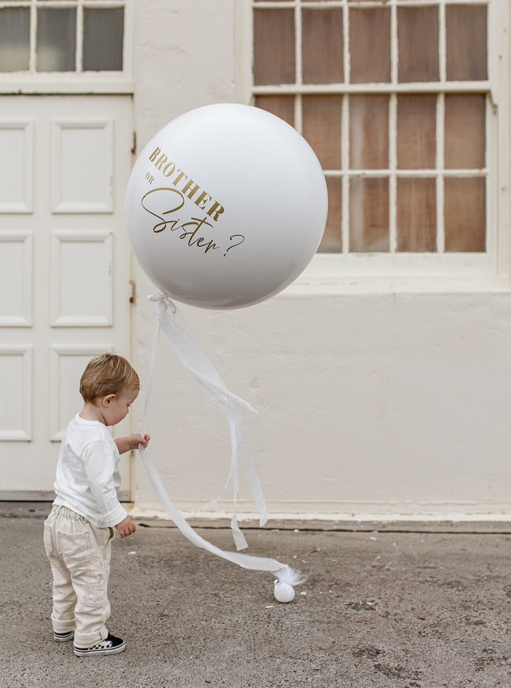 Baby Boy playing with a white gender reveal balloon with custom designed matt gold writing and white faux silk ribbons.