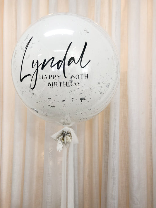 Giant white Birthday helium balloon with silver foil flakes.  The balloon is customised with the birthday girl's name and  message saying  "Happy 60th Birthday" in  black.